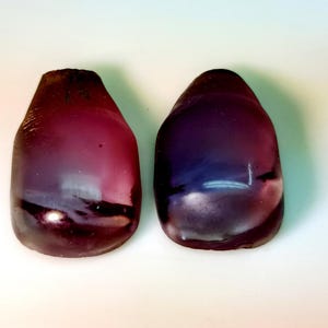 May include: Two smooth, polished, teardrop-shaped cabochons with a deep, translucent purple and dark magenta color gradient. The stones have a glossy, reflective surface with subtle internal variations in tone and light distribution throughout.