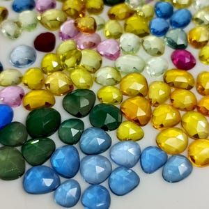 Peut inclure: Une collection de pierres précieuses à facettes de différentes couleurs, dont le bleu, le vert, le jaune, le rose et le rouge. Les pierres précieuses sont de forme ovale et en goutte, avec une surface polie et réfléchissante. Les pierres sont dispersées sur une surface blanche.