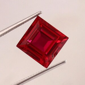 May include: A deep red square-shaped gemstone with a faceted cut. The gemstone is held by a pair of tweezers.