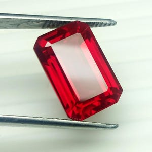 May include: A rectangular, faceted red gemstone held by tweezers. The gem has a clear centre and a polished surface. The edges are sharply defined, showcasing the cut and clarity. The gemstone is a deep, rich red colour.