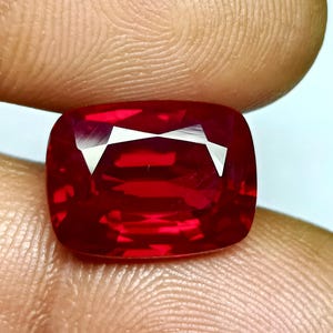May include: A vibrant, cushion-cut ruby gemstone. The gem is a deep, saturated red with a brilliant, reflective surface. The facets create a play of light, highlighting the stone's clarity and color. The ruby is held between fingers.