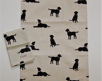 May include: A beige tea towel and folded hand towel featuring a repeating pattern of black dog silhouettes. The folded towel has green stripes and a label that reads "LUCY GOES TO LAPLAND".