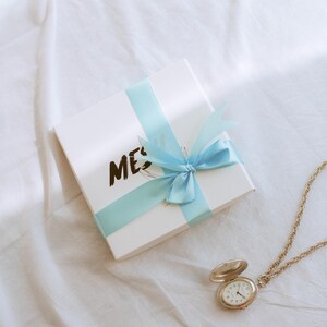 Puede incluir: Una caja de regalo blanca con una cinta azul atada en un lazo. La caja tiene la palabra "ME" escrita en ella. Un reloj de bolsillo dorado con una esfera blanca est&aacute; sobre una superficie blanca.
