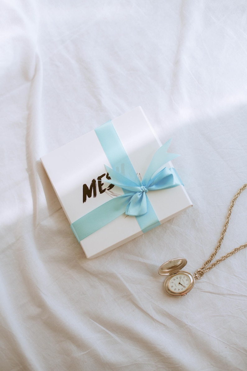 Puede incluir: Una caja de regalo blanca con una cinta azul atada en un lazo. La caja tiene la palabra "ME" escrita en ella. Un reloj de bolsillo dorado con una esfera blanca est&aacute; sobre una superficie blanca.