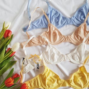 Könnte beinhalten: Vier BHs in verschiedenen Farben: hellblau, beige, weiß und gelb. Jeder BH hat eine zarte Spitzenverzierung und ein Mesh-Etikett mit der Aufschrift "MESH".