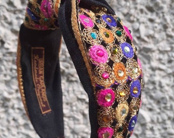May include: Black headband with a knotted design and colorful embroidered embellishments. The headband is decorated with pink, blue, green, and gold beads and sequins. The text "Iran Garlands Handmade in England" is printed on the headband.