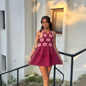 May include: A magenta crochet dress with a halter neck and a flared skirt. The bodice is decorated with white floral motifs. The outfit is completed with woven sandals. The background shows a white building and a set of steps.