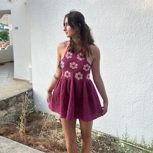May include: A magenta crochet halter dress with white and magenta floral accents. The dress has a flared skirt and a halter neckline. The dress is handmade and suitable for warm weather. The dress is a mini dress.