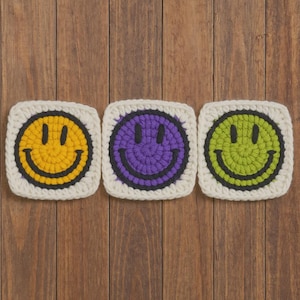 Puede incluir: Tres cuadrados de ganchillo con caras sonrientes. Cada cuadrado tiene un borde blanco y una cara sonriente de diferente color: amarillo, morado y verde. Las caras sonrientes tienen ojos negros y una boca negra curvada. Los cuadrados están dispuestos sobre una superficie de madera.