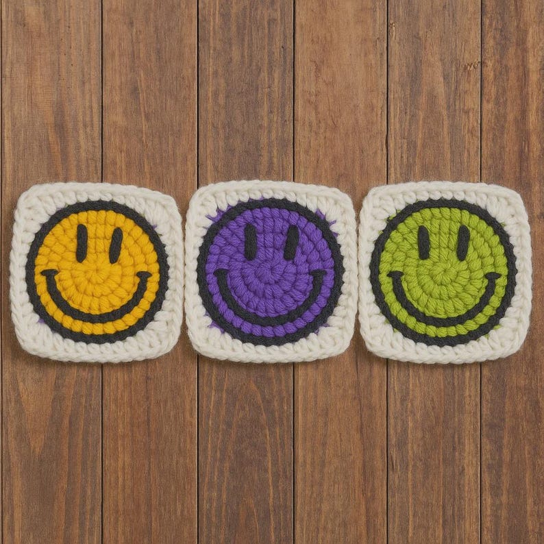 May include: Three crocheted squares featuring smiley faces. Each square has a white border and a different coloured smiley face: yellow, purple, and green. The smiley faces have black eyes and a black curved mouth. The squares are arranged on a wooden surface.