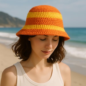 Könnte beinhalten: Ein gehäkelter Bucket Hat mit horizontalen Streifen in Orange und Gelb. Der Hut, getragen von einer Person am Strand, hat eine breite Krempe und eine abgerundete Krone. Der Hut ist aus weichem, strukturiertem Garn gefertigt.