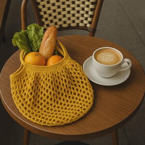 Puede incluir: Una bolsa de mercado amarilla de ganchillo llena de naranjas, lechuga y una baguette est&aacute; sobre una mesa de madera junto a una taza de caf&eacute;. La bolsa tiene un dise&ntilde;o de red. El caf&eacute; est&aacute; en una taza y un platillo blancos. Una silla de madera est&aacute; en el fondo.