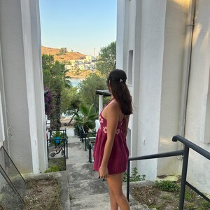 May include: A person wearing a burgundy dress with floral embroidery, standing on concrete steps. The dress has a halter neckline. The person is looking out at a scenic view of a bay and buildings, framed by white walls.