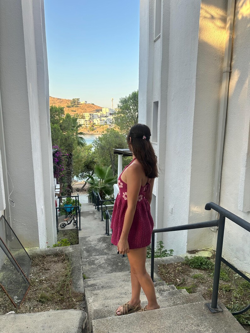 May include: A person wearing a burgundy dress with floral embroidery, standing on concrete steps. The dress has a halter neckline. The person is looking out at a scenic view of a bay and buildings, framed by white walls.