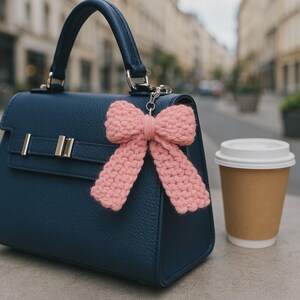 May include: A navy blue handbag with a pink crocheted bow attached. The bag has a top handle and silver hardware. A disposable coffee cup with a white lid sits next to the bag.