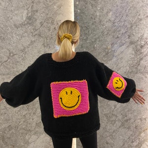 Puede incluir: Su&eacute;ter negro de punto con un parche grande y cuadrado de carita sonriente rosa y amarillo en la parte delantera y uno m&aacute;s peque&ntilde;o en la manga. El su&eacute;ter tiene cuello redondo y mangas largas.