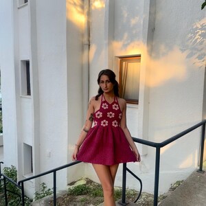 May include: A burgundy halter dress with a floral pattern on the bodice and a flared skirt. The dress is worn with sandals. The background features a white building with a staircase and a black railing.