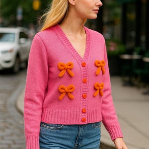 Op de afbeelding: Een felroze vest met oranje strikaccenten en knopen. De V-hals trui wordt gecombineerd met een blauwe spijkerbroek. Het model staat buiten op een stadsstraat.