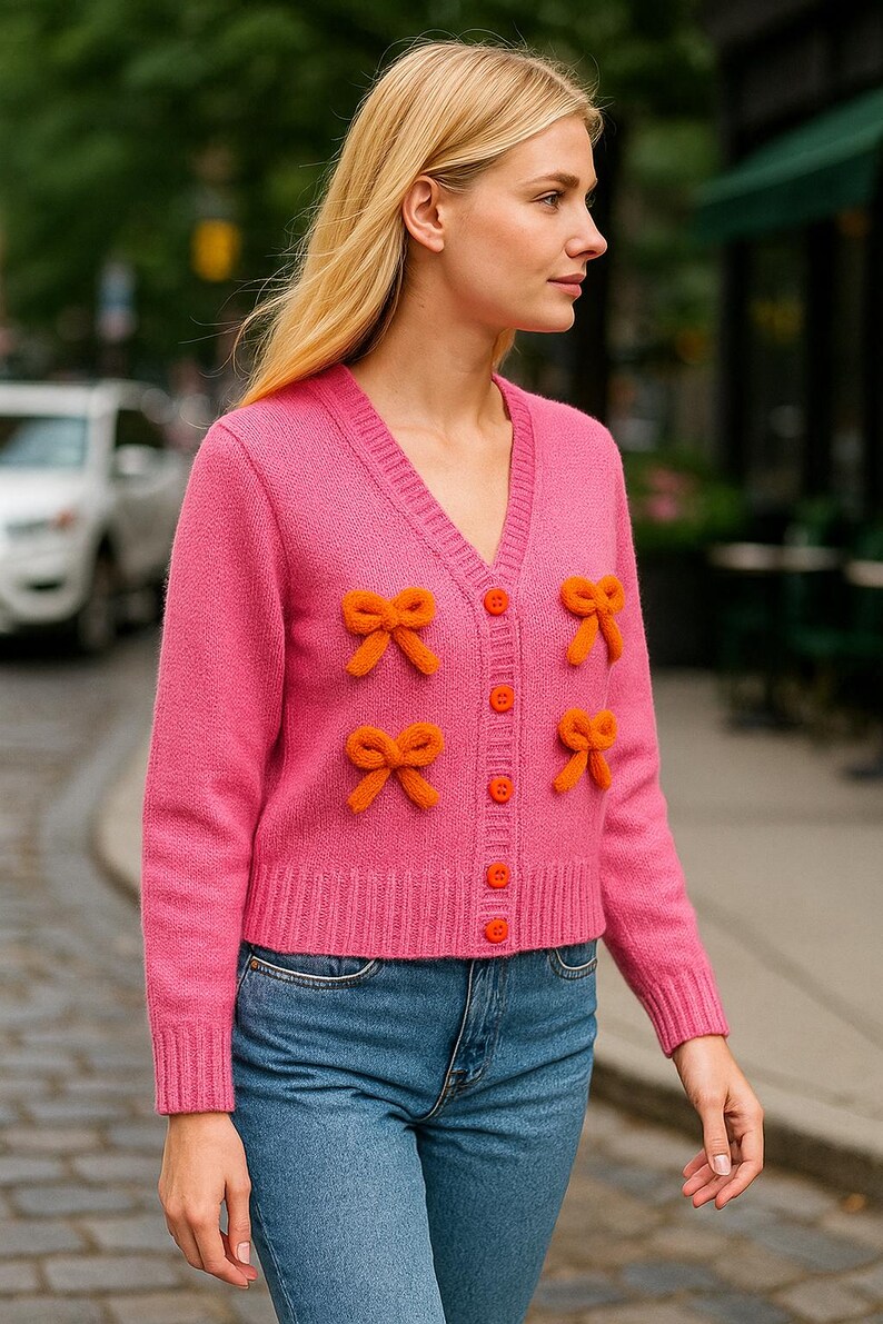 Op de afbeelding: Een felroze vest met oranje strikaccenten en knopen. De V-hals trui wordt gecombineerd met een blauwe spijkerbroek. Het model staat buiten op een stadsstraat.
