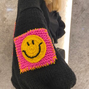Puede incluir: Su&eacute;ter negro de punto con un parche cuadrado grande en la manga. El parche presenta una carita sonriente amarilla sobre un fondo rosa, delineada con costuras amarillas. El su&eacute;ter tiene mangas largas y un corte holgado.