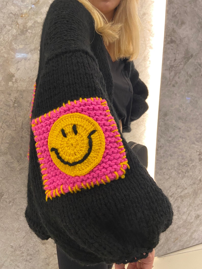 Puede incluir: Su&eacute;ter negro de punto con un parche cuadrado grande en la manga. El parche presenta una carita sonriente amarilla sobre un fondo rosa, delineada con costuras amarillas. El su&eacute;ter tiene mangas largas y un corte holgado.