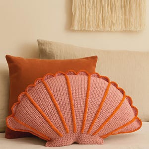 Puede incluir: Un cojín rosa en forma de concha con detalles naranjas. El cojín es de punto y tiene un borde festoneado. Descansa sobre una cama con otros cojines y un edredón estampado. Un tapiz de pared tejido está en el fondo.