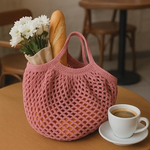 Puede incluir: Una bolsa de red rosa tejida a ganchillo llena de naranjas, una baguette y flores blancas. Una taza de caf&eacute; blanca est&aacute; sobre un platillo junto a la bolsa. La bolsa tiene dos asas y un dise&ntilde;o de malla.