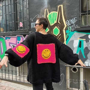 Puede incluir: Su&eacute;ter negro de punto con parches rosas cuadrados grandes con caras sonrientes amarillas. El su&eacute;ter tiene mangas largas y un ajuste relajado. El fondo presenta arte de graffiti en una pared oscura.