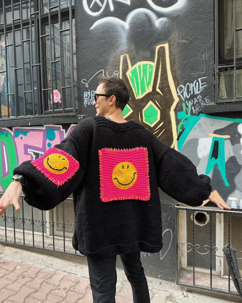 Puede incluir: Su&eacute;ter negro de punto con parches rosas cuadrados grandes con caras sonrientes amarillas. El su&eacute;ter tiene mangas largas y un ajuste relajado. El fondo presenta arte de graffiti en una pared oscura.