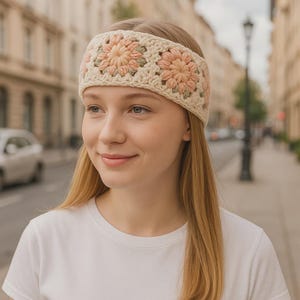 May include: Cream-colored crochet headband featuring three floral designs in peach and green. The headband is worn across the forehead. The model is wearing a white t-shirt.