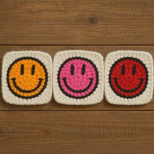 May include: Three crocheted smiley face squares in yellow, pink, and red with black outlines and smiles, set against a cream-coloured background. The squares are arranged on a wooden surface.