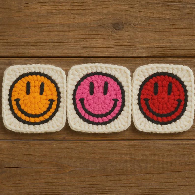 May include: Three crocheted smiley face squares in yellow, pink, and red with black outlines and smiles, set against a cream-coloured background. The squares are arranged on a wooden surface.
