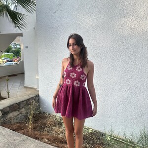May include: A magenta crochet halter dress with a flared skirt. The bodice features white floral accents. The dress is worn with tan sandals. The background is a white textured wall and a concrete walkway.