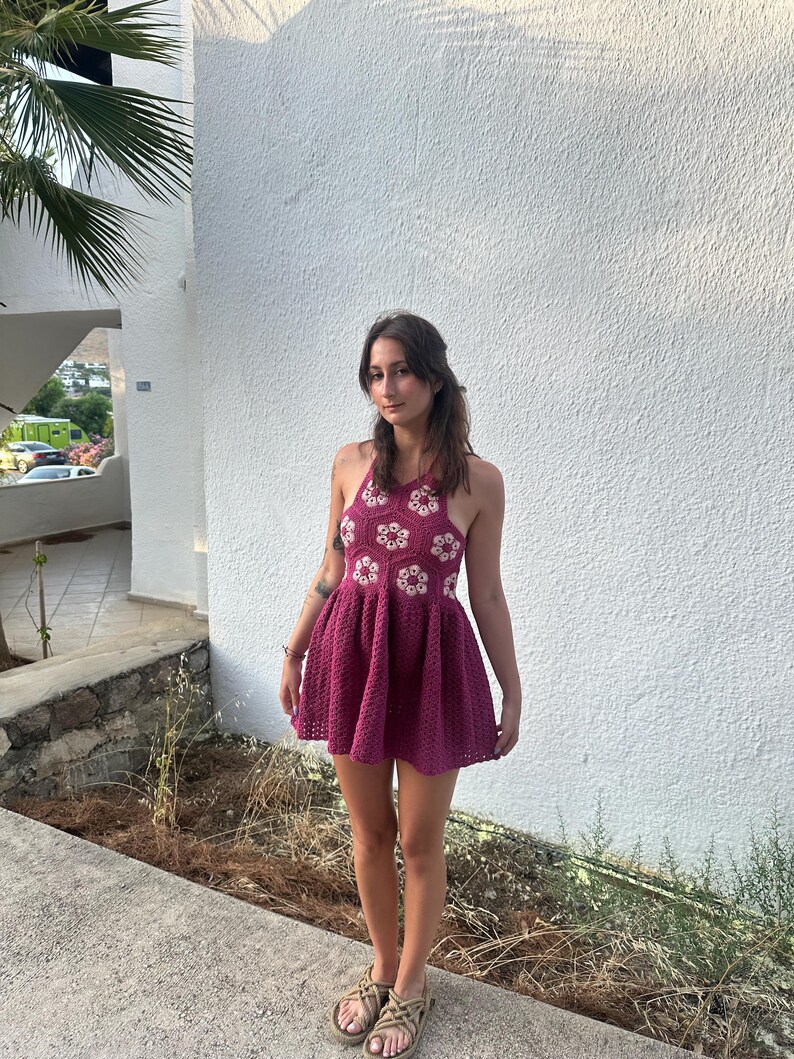 May include: A magenta crochet halter dress with a flared skirt. The bodice features white floral accents. The dress is worn with tan sandals. The background is a white textured wall and a concrete walkway.