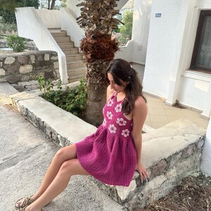 May include: A magenta crochet dress with white floral detailing. The halter-neck dress has a flared skirt and is paired with woven sandals. The background includes a stone wall, stairs, and a palm tree, suggesting a resort setting.