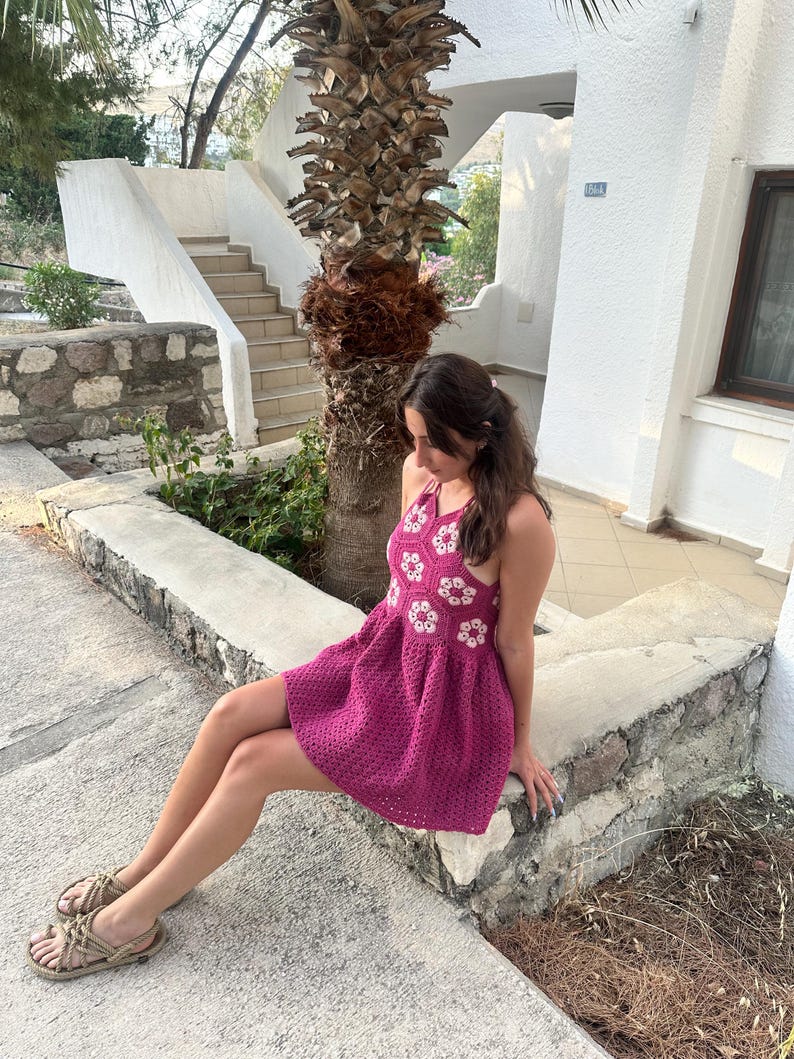 May include: A magenta crochet dress with white floral detailing. The halter-neck dress has a flared skirt and is paired with woven sandals. The background includes a stone wall, stairs, and a palm tree, suggesting a resort setting.