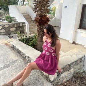 May include: A magenta crochet dress with white floral detailing. The dress has a halter neck and a flared skirt. The person is wearing beige sandals. The background includes a palm tree, steps, and a white building.