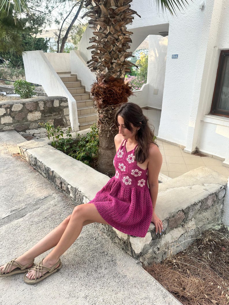 May include: A magenta crochet dress with white floral detailing. The dress has a halter neck and a flared skirt. The person is wearing beige sandals. The background includes a palm tree, steps, and a white building.