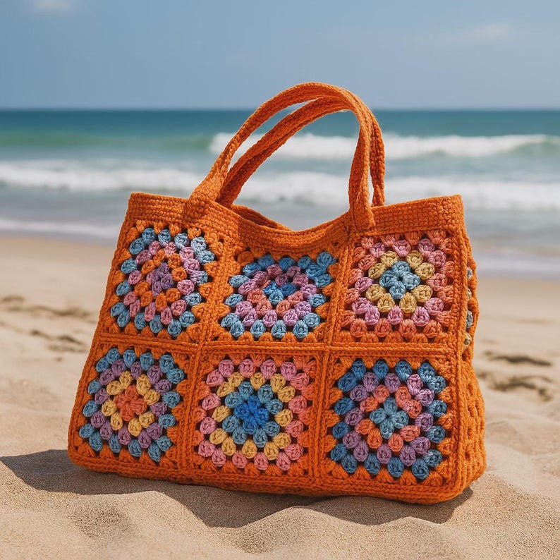 Puede incluir: Un bolso tote de crochet naranja con un colorido estampado floral. El bolso presenta un dise&ntilde;o cuadrado con un patr&oacute;n repetitivo de cuadrados azules, rosas, amarillos y morados. El bolso tiene dos asas y est&aacute; sobre una playa de arena.