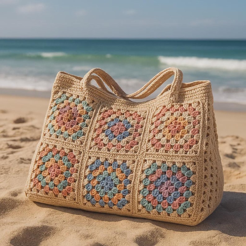 Puede incluir: Un bolso tote de crochet beige con un colorido estampado floral. El bolso est&aacute; hecho de cuadrados con colores pastel, incluyendo rosa, azul y naranja. El bolso tiene dos asas y est&aacute; sobre una playa de arena.