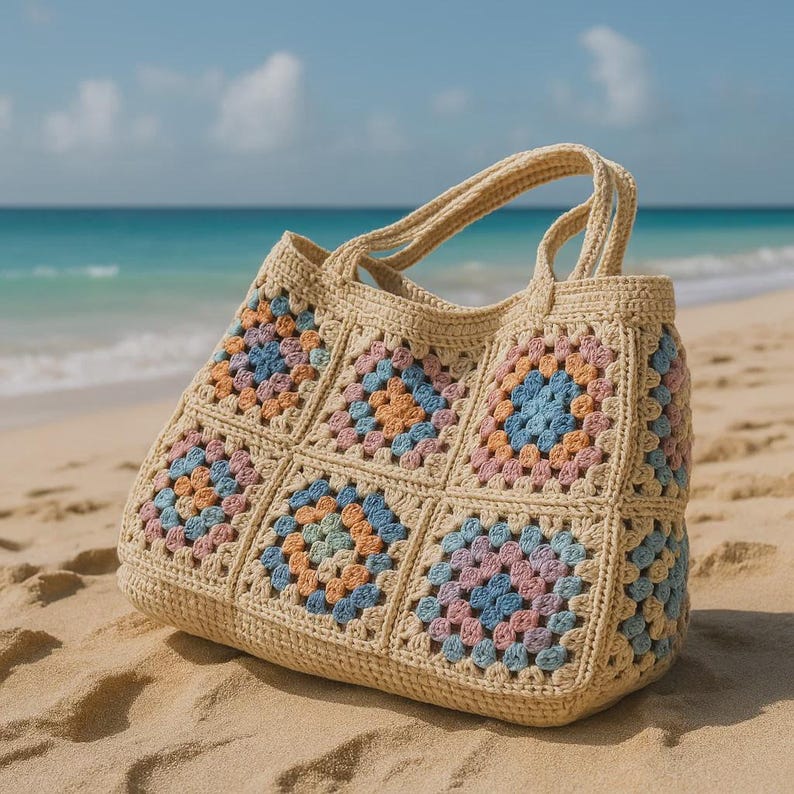 Puede incluir: Un bolso tote de crochet beige con un dise&ntilde;o de cuadrados de la abuela. Los cuadrados presentan colores pastel, incluyendo rosa, azul y naranja. El bolso tiene dos asas y est&aacute; sobre una playa arenosa.