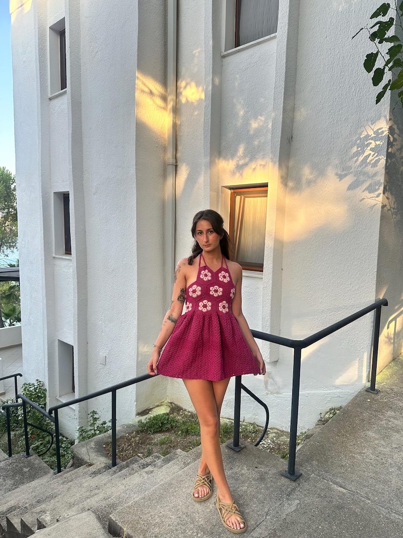 May include: A magenta crochet dress with a halter neck and a flared skirt. The bodice is decorated with white floral motifs. The outfit is completed with woven sandals. The background shows a white building and a set of steps.