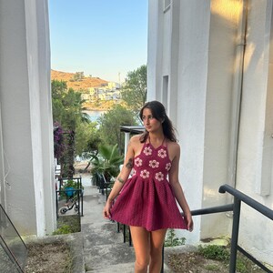 May include: A magenta halter-neck crochet dress. The bodice has a floral design with white and magenta details. The dress has a flared skirt, suitable for summer. The model is wearing sandals. The dress is approximately 80 cm in length.