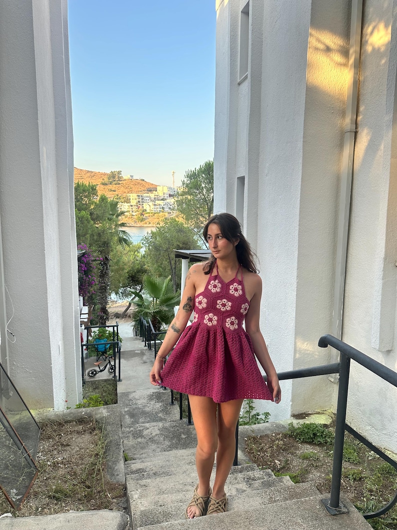 May include: A magenta halter-neck crochet dress. The bodice has a floral design with white and magenta details. The dress has a flared skirt, suitable for summer. The model is wearing sandals. The dress is approximately 80 cm in length.