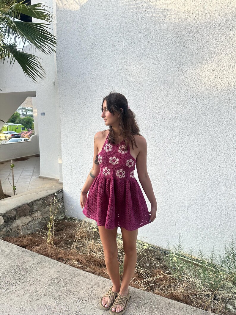 May include: A magenta crochet halter dress with white and magenta floral accents. The dress has a flared skirt and a halter neckline. The dress is handmade and suitable for warm weather. The dress is a mini dress.