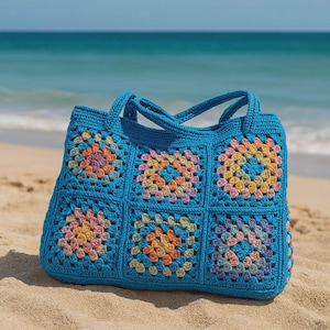 Puede incluir: Un bolso tote de crochet azul hecho a mano con un dise&ntilde;o de cuadrados de la abuela. El bolso presenta seis cuadrados con coloridos dise&ntilde;os florales en tonos rosa, naranja, amarillo y morado. Tiene dos asas y est&aacute; sobre una playa arenosa.