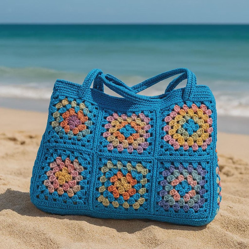 Puede incluir: Un bolso tote de crochet azul hecho a mano con un dise&ntilde;o de cuadrados de la abuela. El bolso presenta seis cuadrados con coloridos dise&ntilde;os florales en tonos rosa, naranja, amarillo y morado. Tiene dos asas y est&aacute; sobre una playa arenosa.