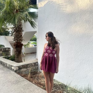 May include: A burgundy crochet halter dress with white flower detailing. The dress features a flared skirt and is paired with tan sandals. The background includes a white wall, palm tree, and a paved walkway.