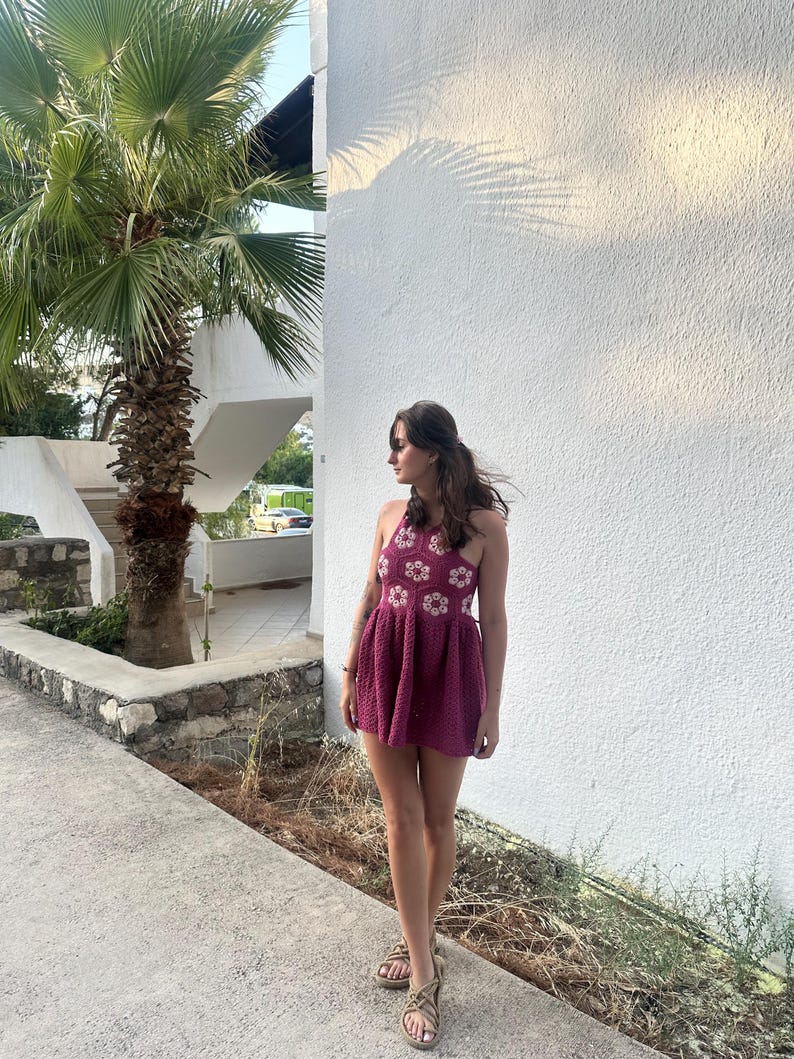 May include: A burgundy crochet halter dress with white flower detailing. The dress features a flared skirt and is paired with tan sandals. The background includes a white wall, palm tree, and a paved walkway.