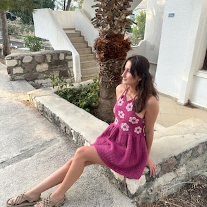 May include: A magenta crochet dress with white floral detailing. The dress has a halter neck and a flared skirt. The image also shows beige sandals with woven straps. The background includes a stone wall, steps, and a palm tree.